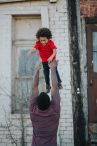 Deeply hued Black man in maroon shirt playfully throwing son in the air.