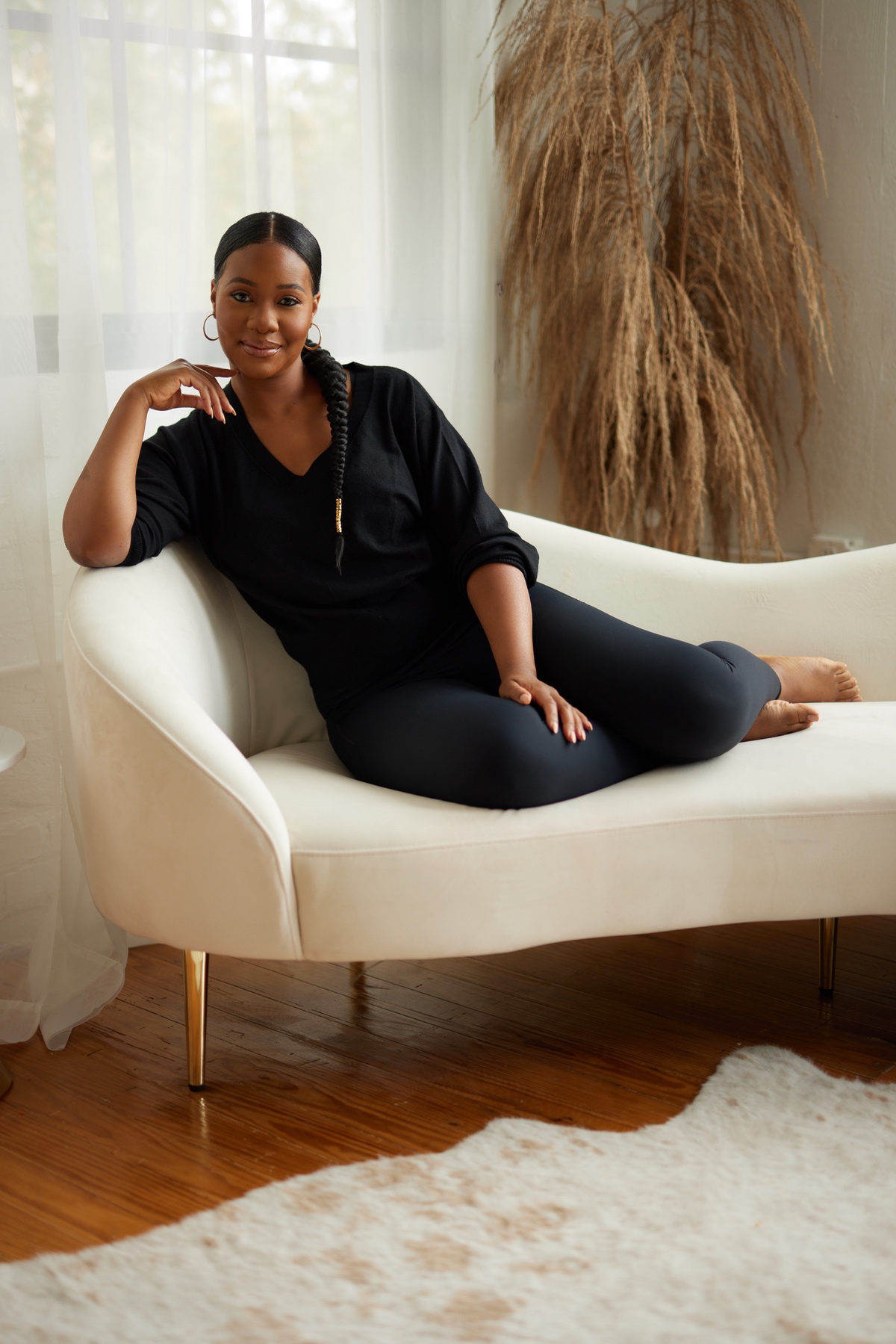Jasmine Marie is a Black woman with a long braided hairstyle sits comfortably on a white curved chaise lounge or modern sofa. She's wearing all-black attire consisting of a V-neck top and fitted pants. Her posture is relaxed yet poised as she rests against the sofa with one hand thoughtfully positioned near her face. The serene setting includes soft natural light coming through sheer curtains, a wooden floor, a partial view of a white area rug, and decorative dried pampas grass in the background. The overall aesthetic is calm, minimalist and wellness-focused.